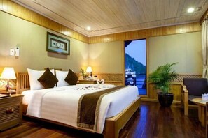 Une chambre d’hôtel avec un lit en bois, des tables de chevet, une plante en pot et une vue sur une montagne, visible à travers une grande fenêtre.
