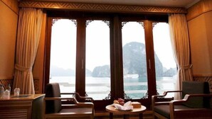 A room with a view of a body of water and a mountain, featuring wooden furniture and curtains. - Pelican Classic Cruise (Tuan Chau)