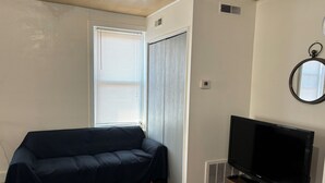 Ein Wohnzimmer mit einem dunkelgrauen Sofa, einem Flachbildfernseher auf einem Holzboden und einem Fenster mit Jalousien. An der Wand ist eine Klimaanlage angebracht.