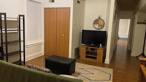 Ein Wohnzimmer mit Fernseher, einem Holzschrank, einem schwarzen Ottomanen und einem gemusterten Teppich.