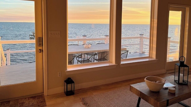 A room with three large windows overlooking a dock and the sea. There is a wooden table with a bowl and a lantern. - Private Waterfront Beach House with Fire-pit (Ocean Springs)