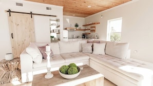 A modern living room with a large L-shaped sectional sofa, a wooden coffee table with a green decorative bowl, and a kitchen area visible in the background. - Private Waterfront Beach House with Fire-pit (Ocean Springs)