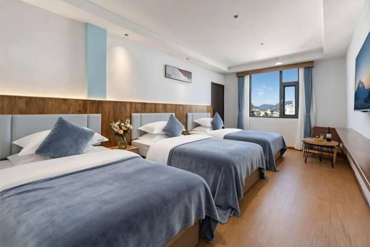 A hotel room with three beds, a wooden headboard, a small table with a bottle, and a view of the city.