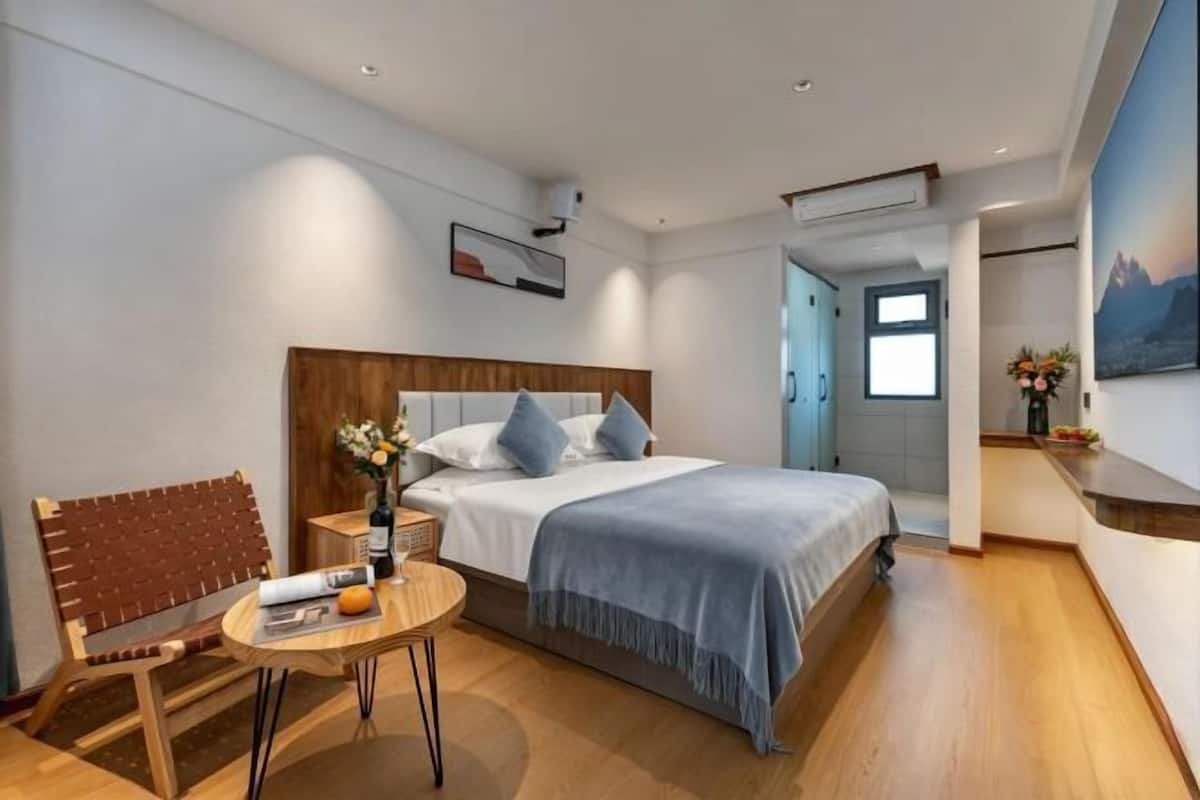 A modern hotel room with a wooden bed, a small round table with a chair, a nightstand, and a painting on the wall.