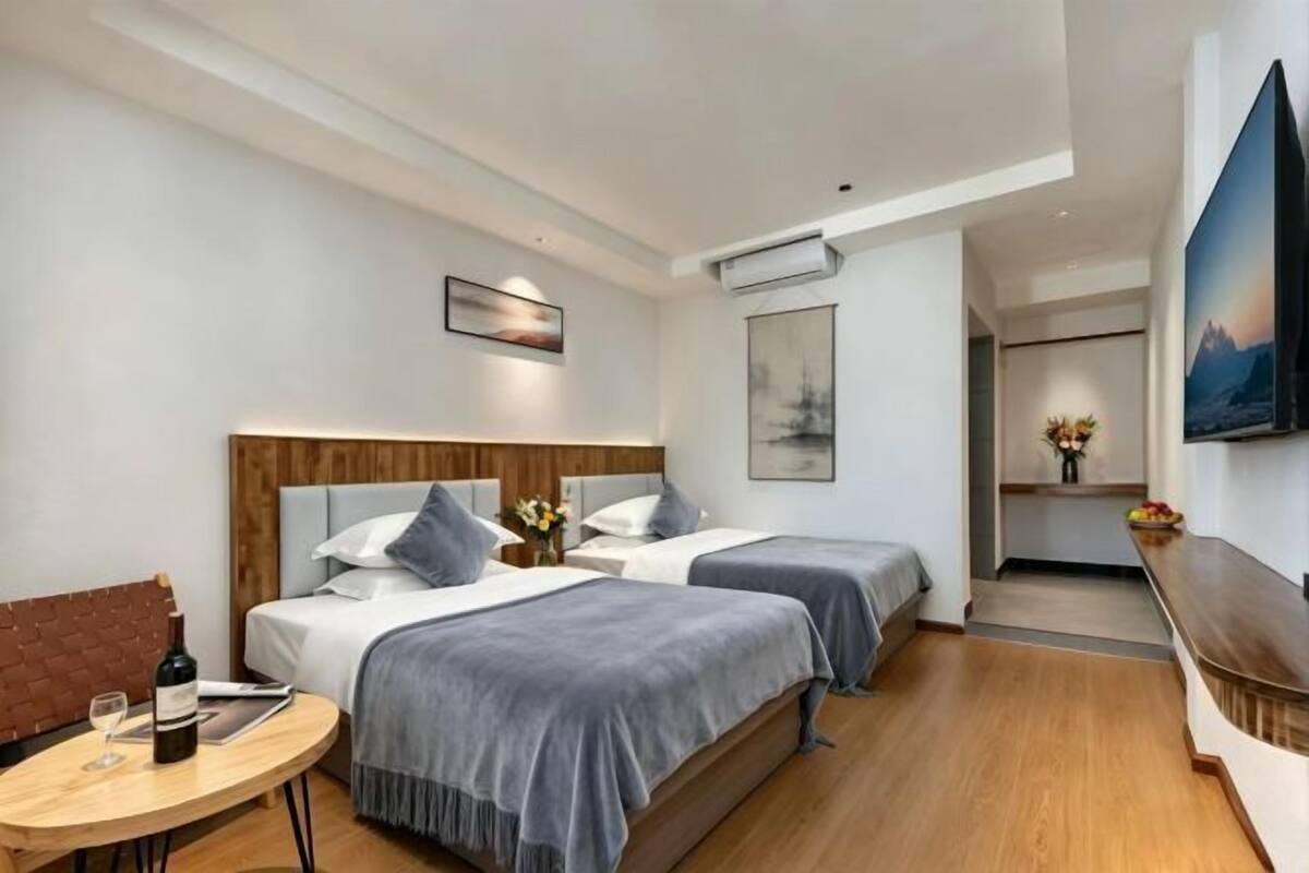 A hotel room with two beds, a small table with a bottle and glass, a TV, and a painting on the wall.