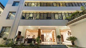Entrada moderna de um hotel com fachada de vidro, área de recepção e escadaria que leva aos andares superiores.