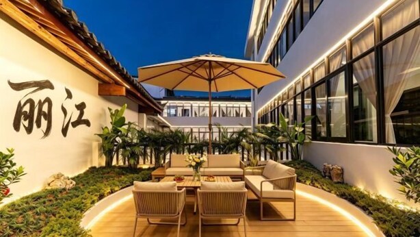 Uma área externa com mesas, cadeiras e um guarda-sol, cercada por plantas e fachada de um edifício com grandes janelas.