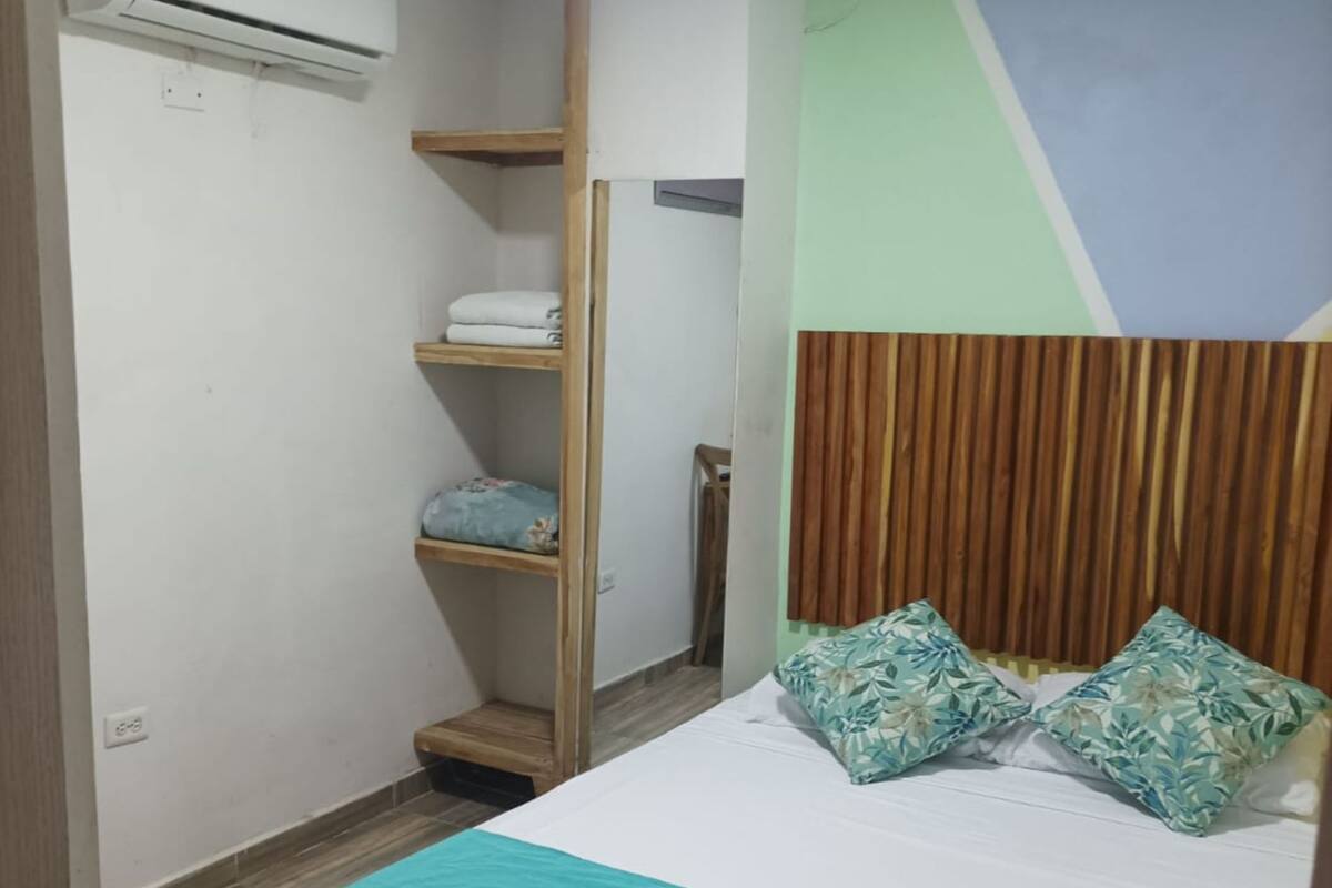 A bedroom with a bed, a wooden headboard, a wall-mounted air conditioner, and a shelf with folded towels.