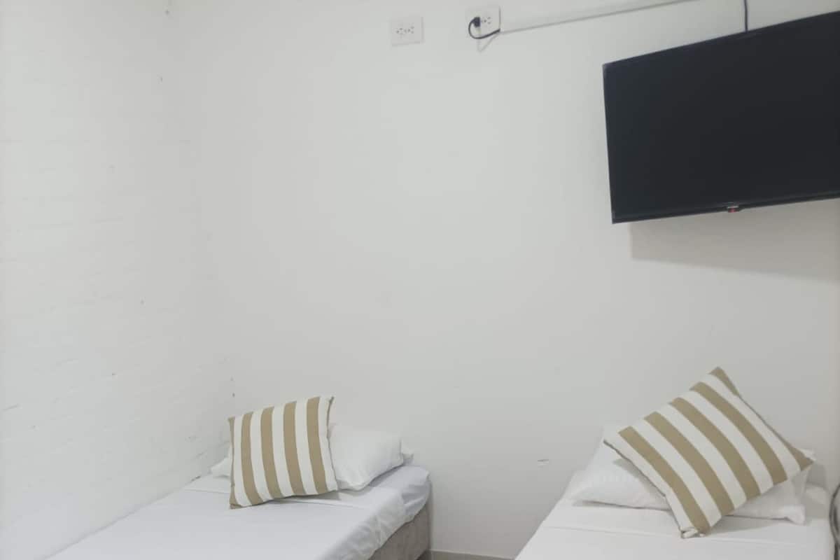 A small, clean room with two single beds, each with a pillow and a white bedspread. A flat-screen TV is mounted on the wall above the beds.