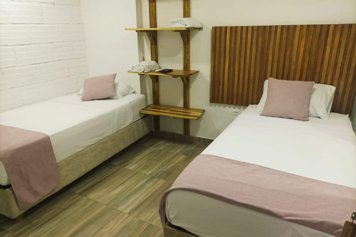 A room with two single beds, each with a pink blanket and pillow. There is a wooden shelf with a white towel and a remote control. The room has a wooden floor and a brown headboard.