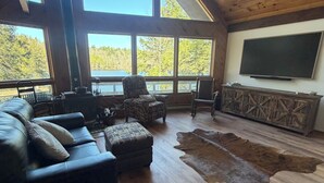 A spacious living room with a large sofa, ottomans, a wooden coffee table, a television, and a large window overlooking a forested area. - Beautiful lakefront house on Paradox lake with tremendous views and privacy. (Paradox)