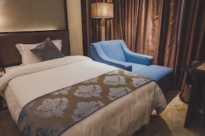 A hotel room with a bed, a blue armchair, a lamp, and a curtain.