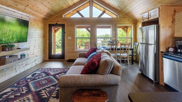 Un chalet confortable avec un intérieur en bois, un canapé avec des coussins, un téléviseur à écran plat, un réfrigérateur et un coin repas avec une table et des chaises.