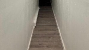 Un pasillo estrecho, tenuemente iluminado, con piso de madera y paredes blancas.