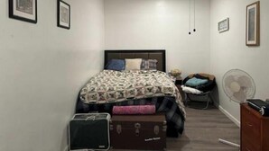 Un dormitorio pequeño y ordenado con cama, una silla, un ventilador y un aparador.
