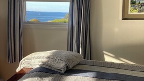 A bedroom with a bed, striped curtains, a window with a view of the sea, and a framed picture on the wall. - Whale Ledge Cottage - In the heart of Lubec with AMAZING Waterviews!  (Lubec)