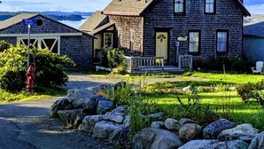A wooden house with a gabled roof, a front porch, and a small garden with rocks and greenery. - Whale Ledge Cottage - In the heart of Lubec with AMAZING Waterviews!  (Lubec)