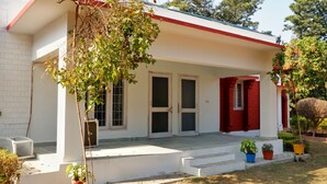 Un edificio con fachada blanca, detalles en rojo y un porche con escalones que conducen a una puerta. Hay plantas en macetas en el porche y un árbol de hojas verdes.
