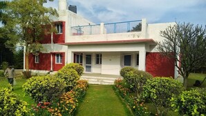 Un edificio de dos pisos con fachada roja y blanca, un balcón y un jardín bien cuidado con diversas flores.