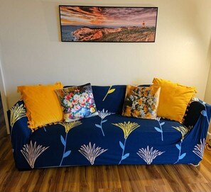 A blue sofa with yellow cushions and floral-patterned pillows, set against a white wall with a framed landscape painting above.
