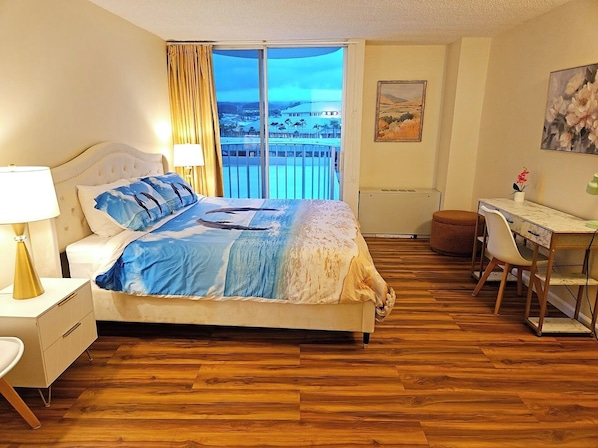 A bedroom with a bed, a desk, a chair, a lamp, and a view of a cityscape through a large window.