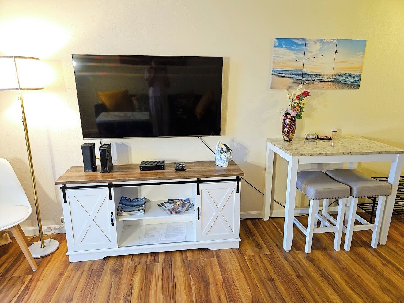 A room with a TV mounted on the wall, a wooden cabinet with storage, a small table with two stools, and a lamp.