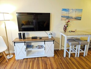 A room with a TV mounted on the wall, a wooden cabinet with storage, a small table with two stools, and a lamp. - Newly Renovated Hilo Lagoon 2 BD Condo-A/C (Hilo)