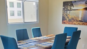 A dining area with a wooden table and blue chairs, a window with blinds, a painting of a sunset, and a hanging light fixture.
