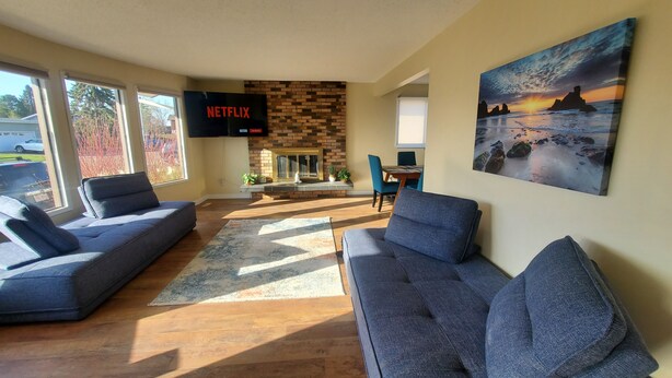 A modern living room with two grey sofas, a fireplace, a flat-screen TV displaying 'NETFLIX', and a large painting of a sunset over the ocean.
