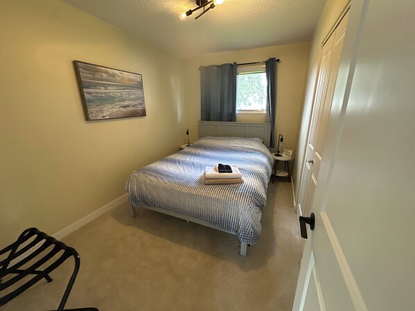 A bedroom with a bed, a nightstand, a lamp, a window with curtains, a painting on the wall, and a chair.
