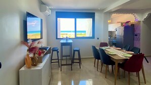 A spacious living area with a large window offering a view of the sea, a dining table set for four, and a kitchen area visible through an archway. - Bel Appartement Moderne , Vue Océan Front de Mer, Plage, Nature, Détente (Tanger)