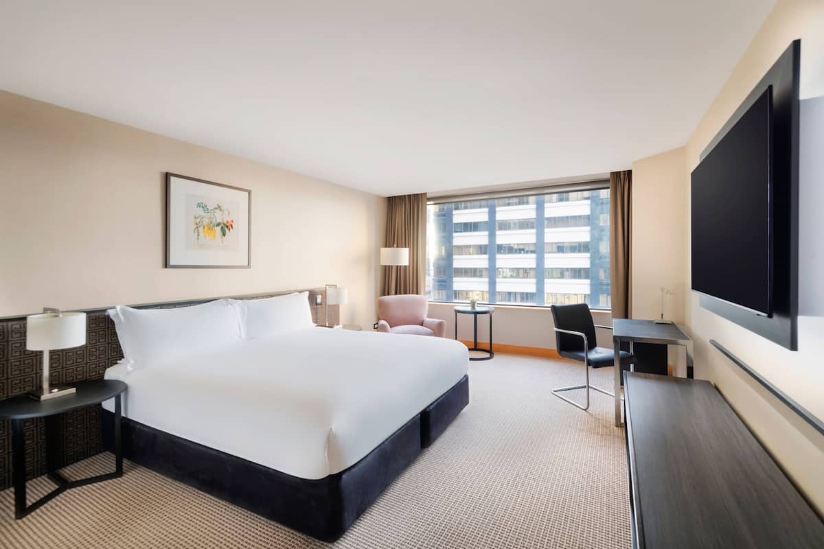Quarto de hotel com uma cama grande, uma escrivaninha, uma cadeira, televisão e janela com vista para a cidade.