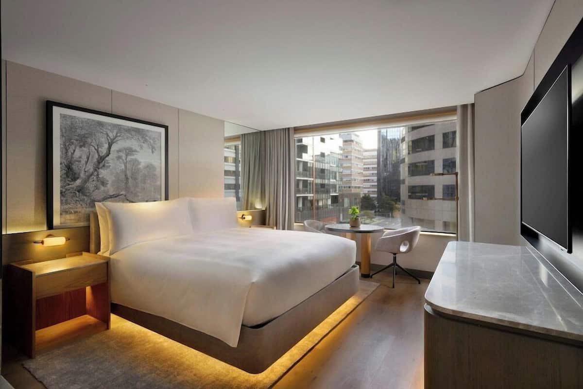Quarto de hotel moderno com uma cama grande, mesas de cabeceira, televisão, uma pequena mesa com cadeiras e vista para a cidade através de janelas amplas.
