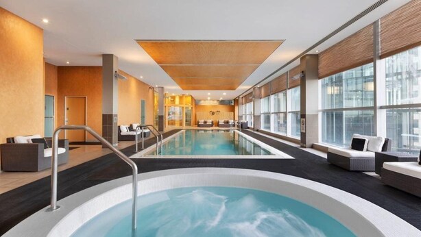A modern indoor swimming pool with a central hot tub, surrounded by seating areas and large windows. - JW Marriott Auckland (Auckland)