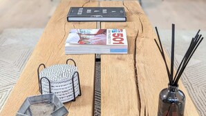 Une table en bois, avec un magazine, un petit sac, un récipient en filet et un diffuseur de parfum à roseaux.