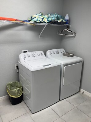 A laundry area with two front-loading washing machines, a shelf with folded clothes, a bottle of detergent, and a trash bin.