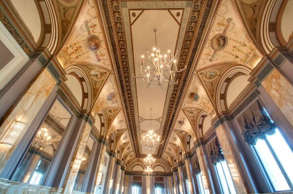 A grand hall with ornate ceiling decorations, a central chandelier, and tall columns.