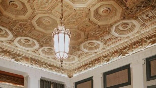 Un vaste salon doté d’un plafond haut et orné de motifs complexes, d’un canapé en cuir marron, d’un fauteuil bleu et d’une table basse.