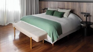Un dormitorio moderno con suelo de madera, una cama con colcha verde y blanca, un banco al pie de la cama y una mesita de noche con una lámpara.