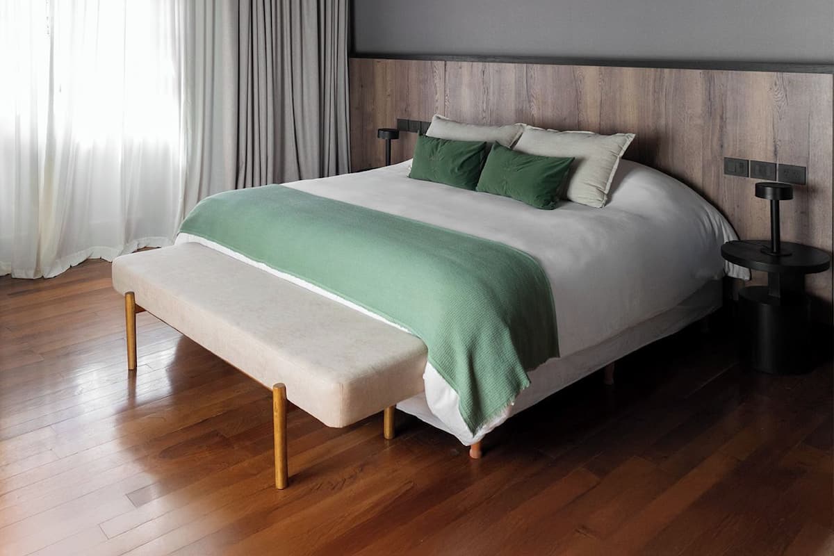 A modern bedroom with a wooden floor, a bed with a green and white bedspread, a bench at the foot of the bed, and a nightstand with a lamp.