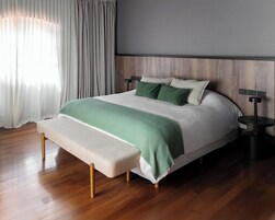 Un dormitorio moderno con suelo de madera, una cama con colcha verde y blanca, un banco al pie de la cama y una mesita de noche con una lámpara.