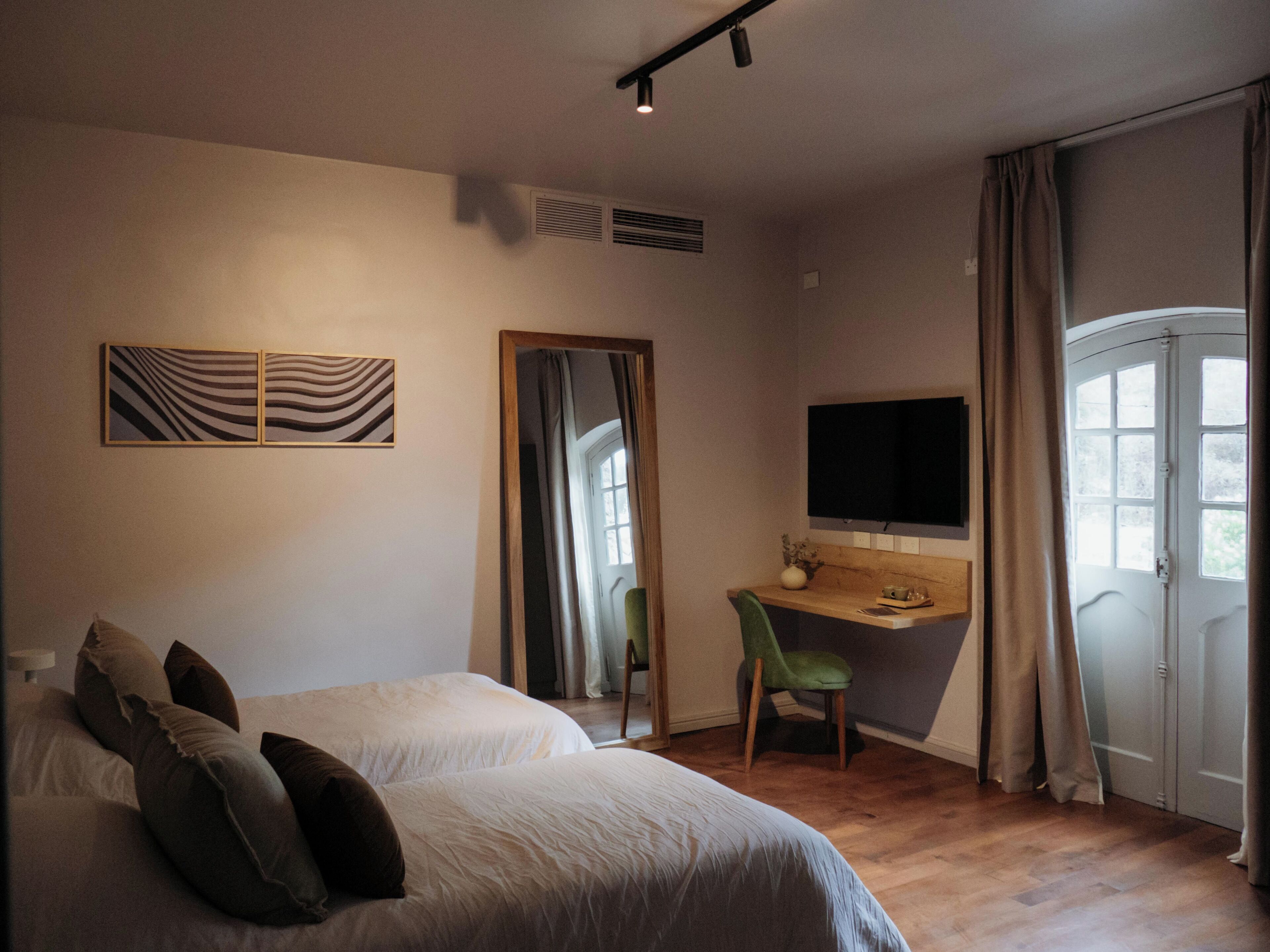 A bedroom with two beds, a desk with a chair, a TV, and a window with curtains.