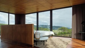 Un dormitorio moderno con una cama grande, un armario de madera, una mesita de noche y vistas a colinas verdes a través de amplias ventanas.