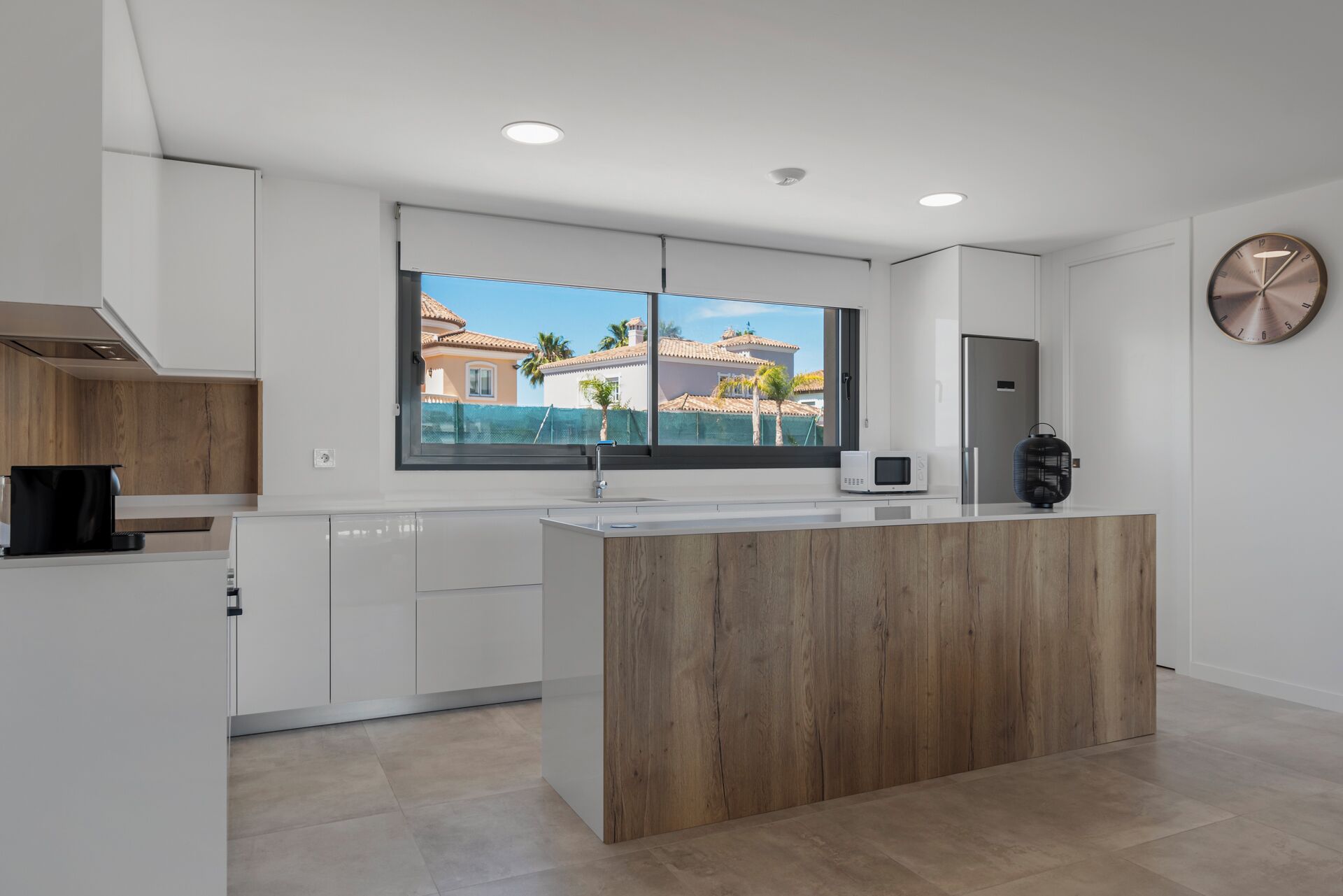 A modern kitchen with a wooden island, white cabinets, a large window with a view of buildings, and a round wall clock.