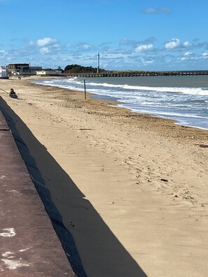 Une plage avec un rivage sablonneux, une jetée en arrière-plan et une personne assise sur le sable.