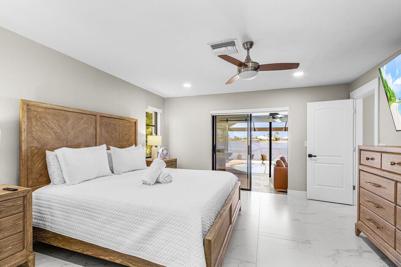 Un dormitorio con una cama grande, cabecera de madera, ventilador de techo y vista a una piscina a través de un ventanal corredero.