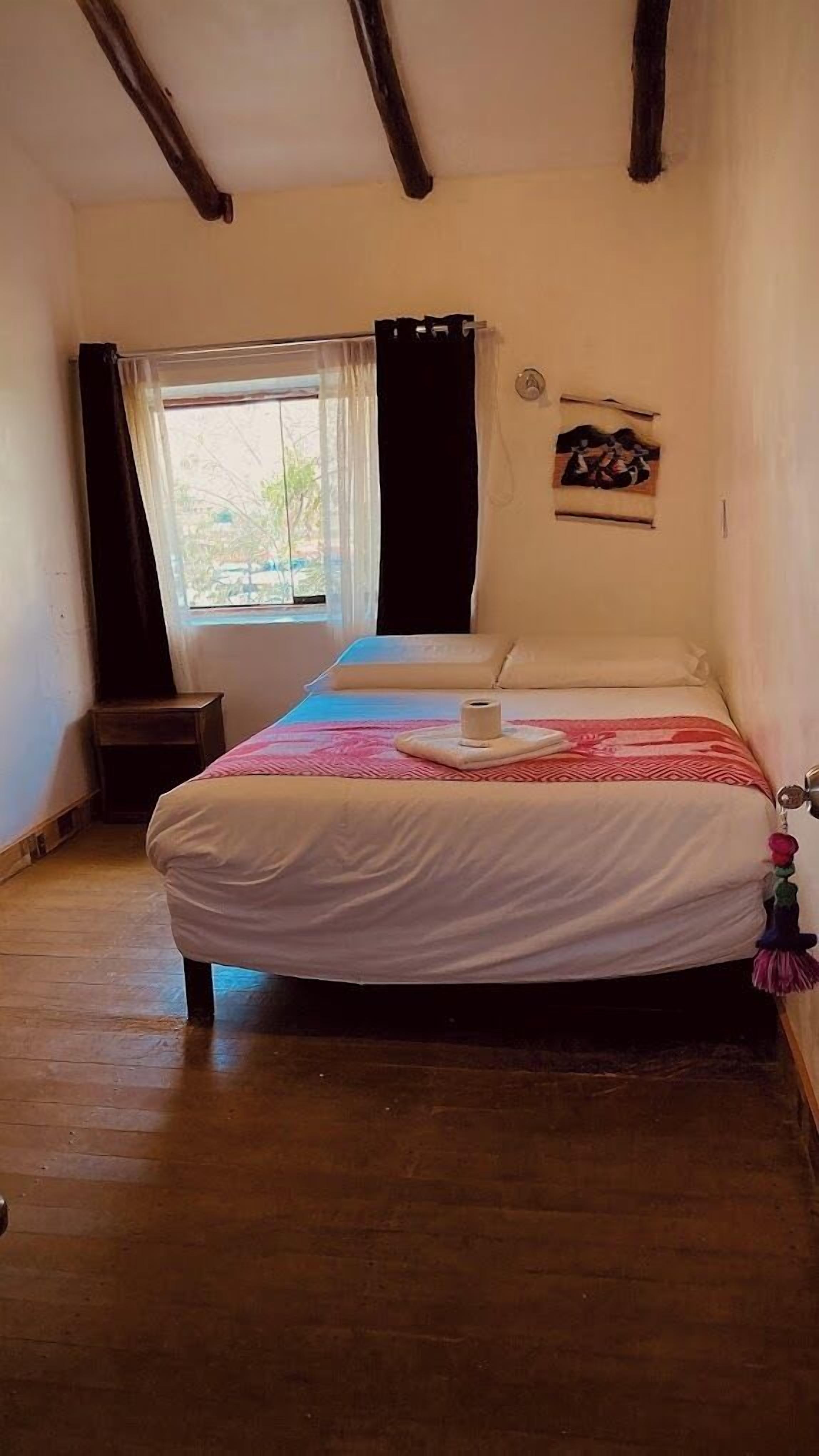 Un lit simple avec un drap blanc et une couverture rose, une petite table de chevet, une fenêtre avec un rideau et un luminaire fixé au mur.