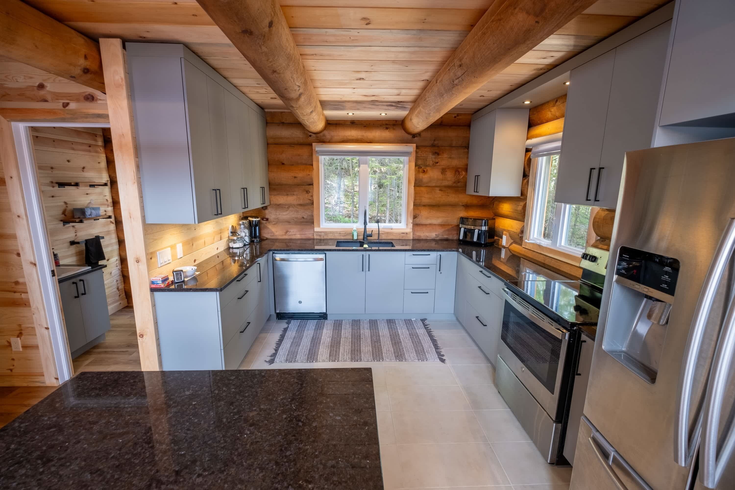 A modern kitchen with wooden beams, grey cabinets, stainless steel appliances, and a large window.
