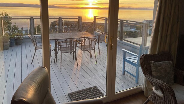 A balcony with a table and chairs, overlooking a sunset over a body of water.