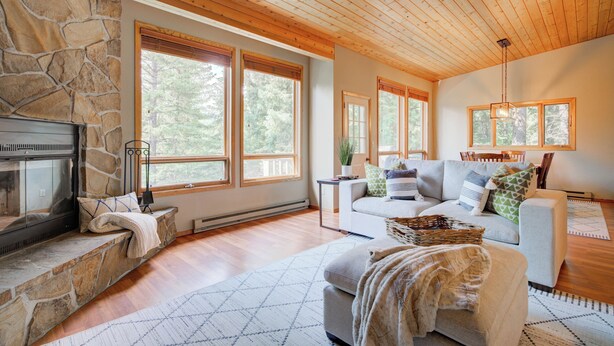 A cozy living room with a stone fireplace, large windows, a white sofa, and a wooden ceiling.
